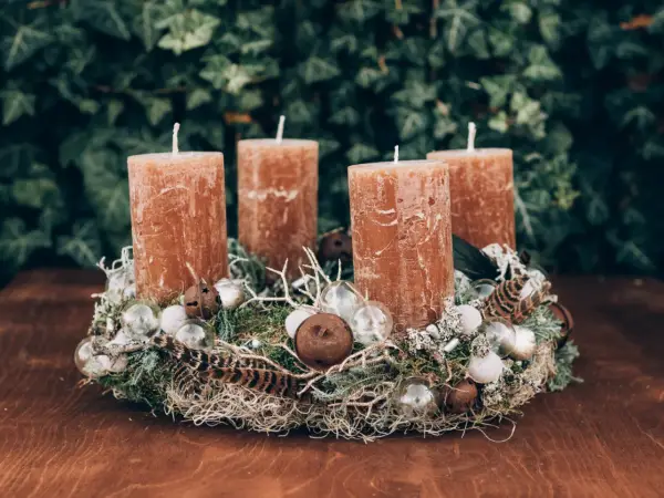 A rustic Advent wreath with four brown candles, moss, feathers, and glass ornaments on a wooden table.